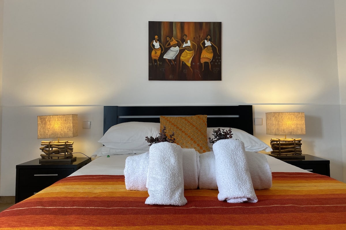 Een hotelkamer met een bed, twee nachtkastjes met lampen, een schilderij aan de muur en een gestreept vloerkleed.