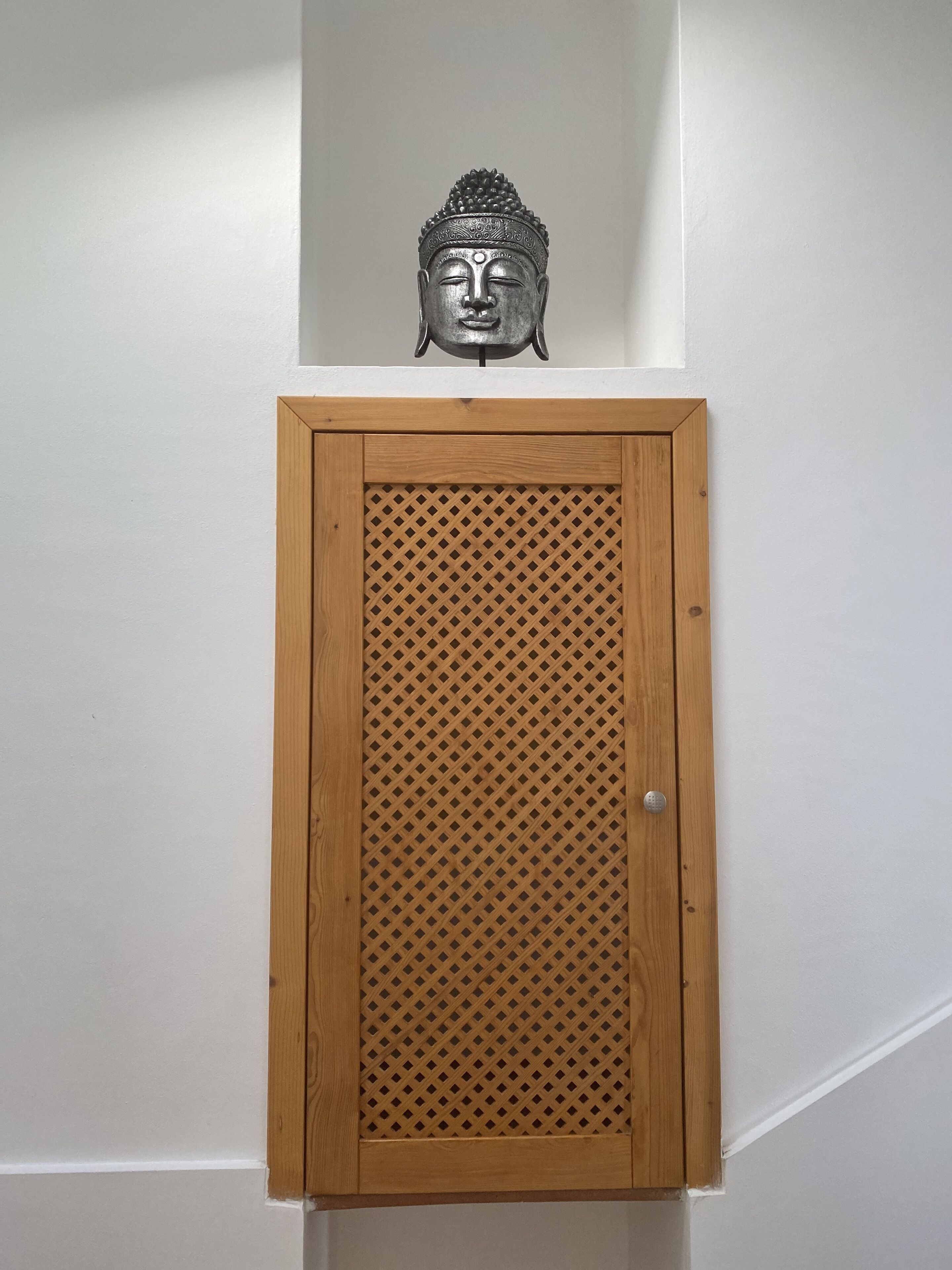 Un mueble de madera con un panel frontal de diseño geométrico en forma de rombos, fijado a una pared blanca sobre una puerta.