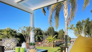 Un patio al aire libre con una mesa de madera, un sofá blanco con un cojín amarillo y diversas macetas con plantas.