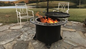 Een vuurplaats met een zwarte waterkoker en stoelen, gelegen op stenen tegels met uitzicht op een meer en bos.