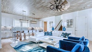 Un salon moderne avec un canapé blanc, des fauteuils bleus et une table basse en verre. On y trouve également un coin cuisine avec des meubles blancs et un bar.