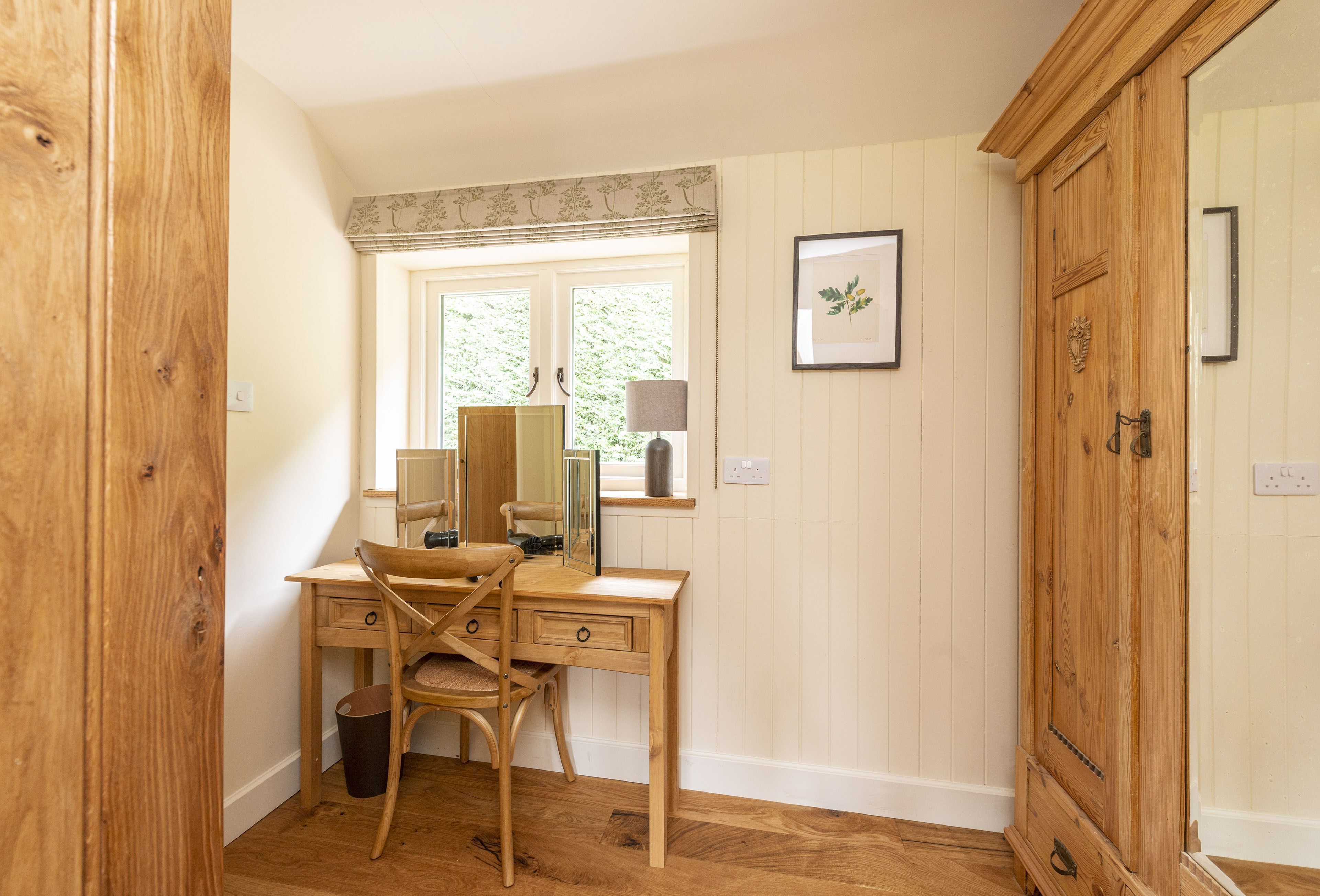 A wooden desk with a chair, a window with blinds, a framed picture on the wall, and a wooden wardrobe.