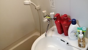 Une salle de bain avec un lavabo blanc, une douchette à main et divers produits de toilette, notamment des flacons rouges et un flacon de shampoing.