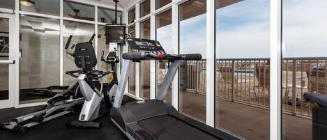 Un gimnasio con caminadora, elíptica y bicicleta estática, ventanas amplias que dan a una piscina y zona exterior.
