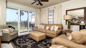 A spacious living room with a beige sectional sofa, a round coffee table, and a black patterned rug. There is a sliding glass door leading to a balcony with a view of palm trees and a building. - Summer Place #109 Large 4Bd Poolside Beachfront - 2 Sets of Beach Service! (Fort Walton Beach)