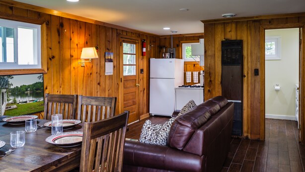 L’intérieur d’une cabane comprend un coin repas, un salon avec un canapé en cuir, des murs en bois, un réfrigérateur et une fenêtre donnant sur un lac.