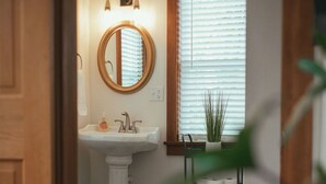 A bathroom with a pedestal sink, a round mirror, and a window with blinds. - Beautiful and Historic Log Cabin: cozy, quiet, huge hot tub! Quiet getaway! (Goshen)