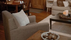 A cozy living room with a herringbone-patterned armchair, a wooden coffee table with a decorative bowl, and a dining area with wooden chairs and a table. - Beautiful and Historic Log Cabin: cozy, quiet, huge hot tub! Quiet getaway! (Goshen)