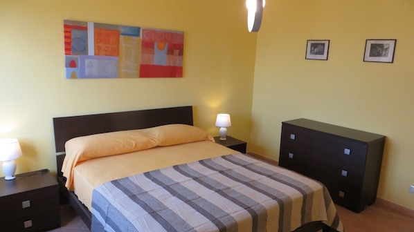 A hotel room with a bed, a nightstand with a lamp, a dresser, and two framed pictures on the wall.