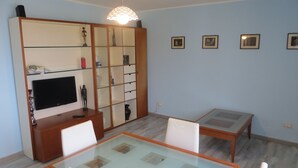 A living room with a TV, glass-top dining table, chairs, a cabinet with glass doors, and a hanging light fixture. - Casa Martina Appartamento Panoramico (Terracina)