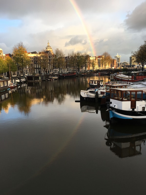 A canal with boats docked, a rainbow in the sky, and buildings in the background.
