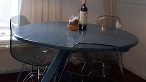 Un coin repas avec une table ronde, des chaises, une lampe suspendue, une bouteille de vin et un sac de pain.
