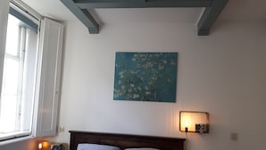 Une chambre à coucher comprenant un lit, une table de chevet, une lampe fixée au mur et une fenêtre avec des stores.
