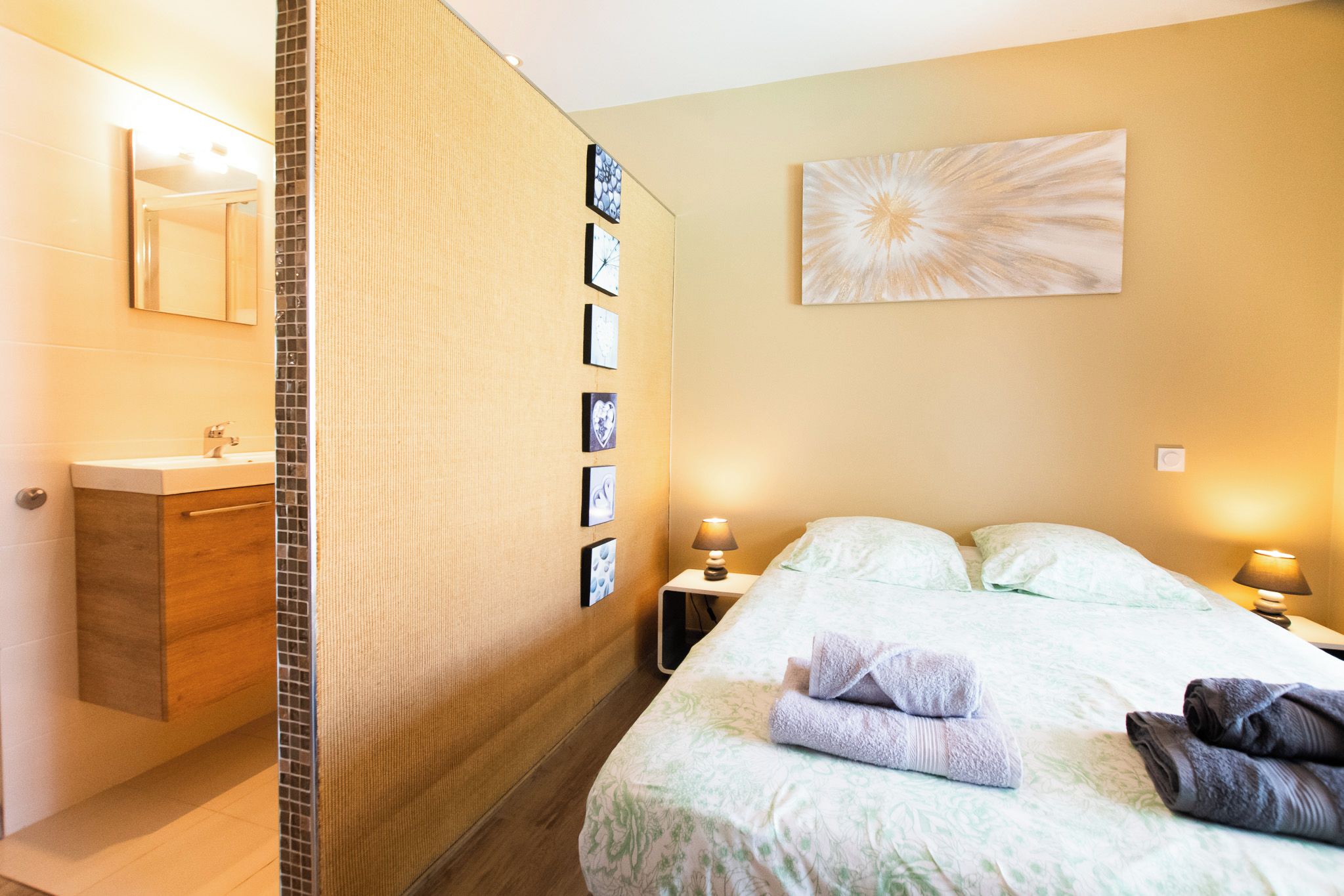 Habitación de hotel con baño, cama con toallas, mesita de noche con lámpara y pared con cuadros enmarcados.