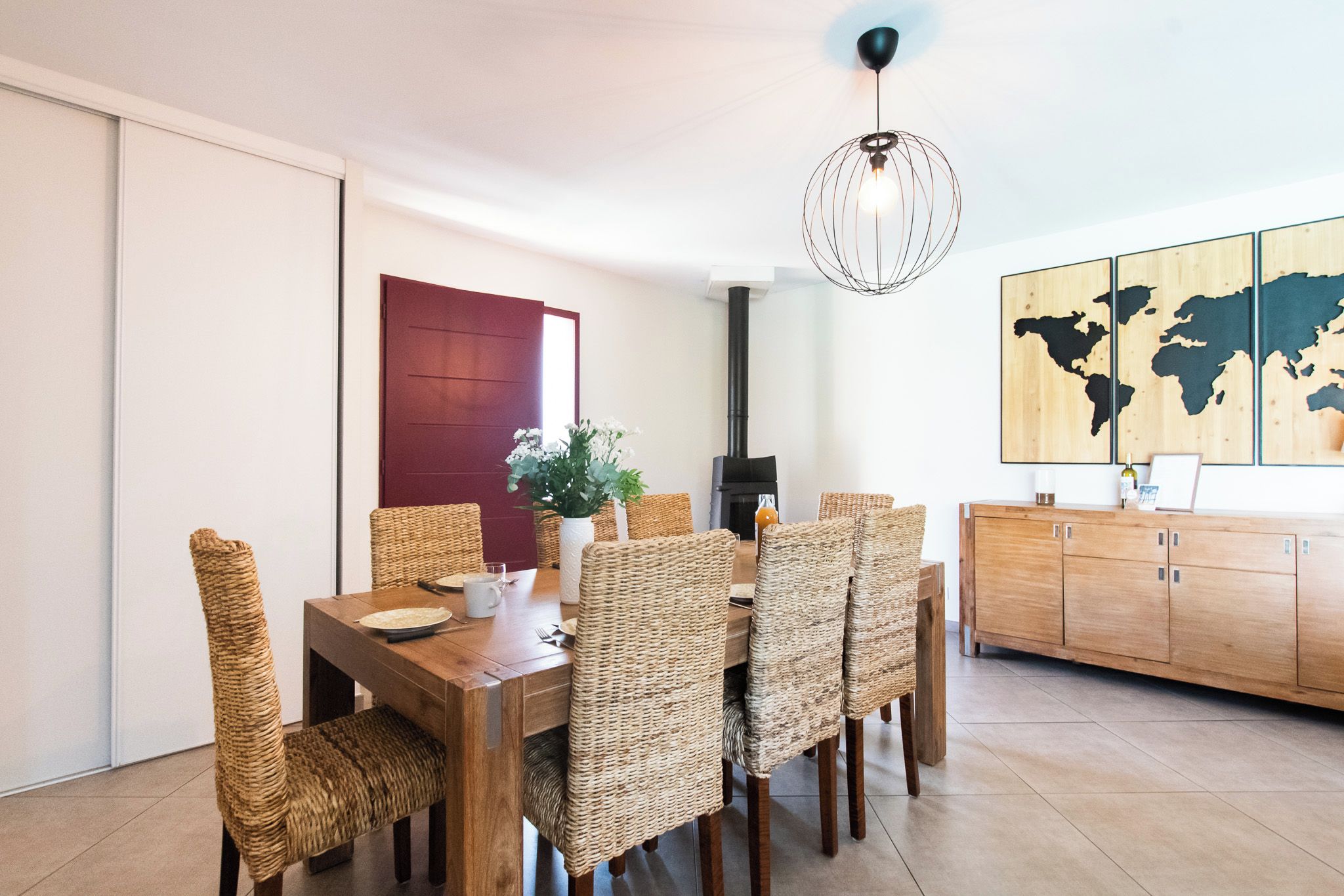 Un comedor con mesa de madera y sillas de mimbre, una puerta roja, una estufa negra y un mapa mundial en la pared.