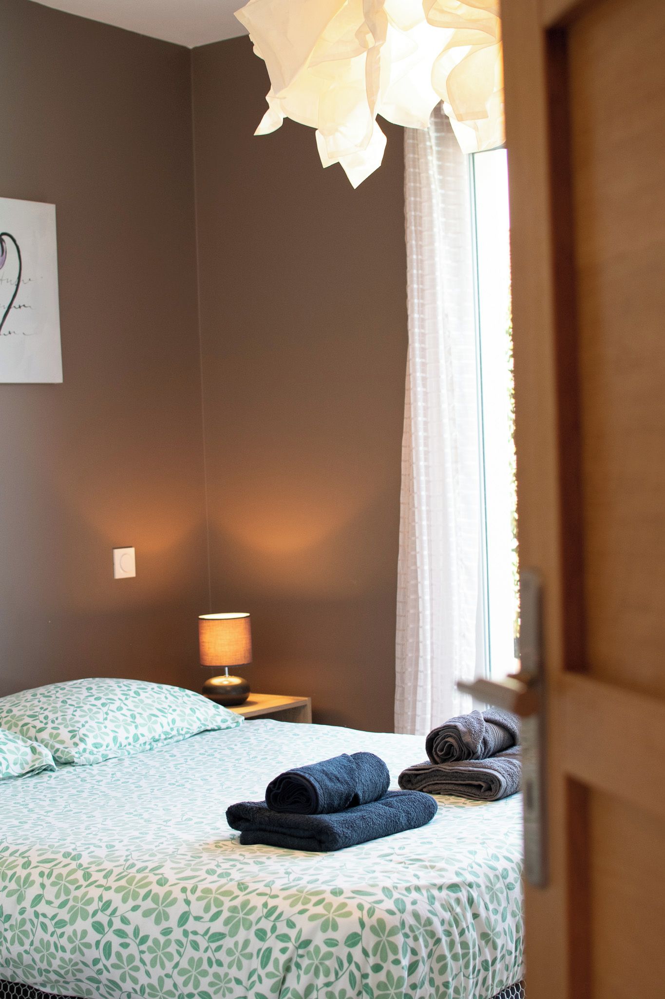 Un dormitorio con cama, mesita de noche, lámpara y ventana con cortinas.