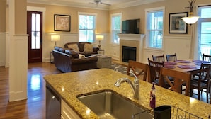 A spacious living room with a fireplace, a dining area with a table and chairs, a kitchen with granite countertops and a sink, and a ceiling fan.