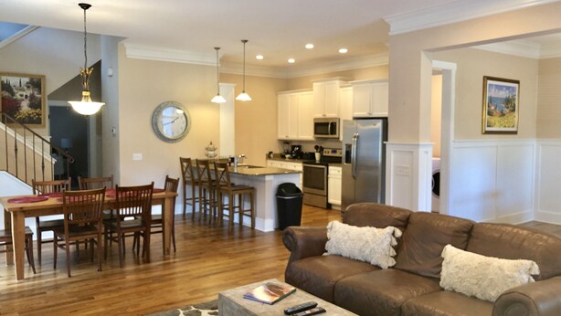 A modern living room with a brown leather sofa, wooden dining table, and chairs. There is a kitchen area with white cabinets and stainless steel appliances.