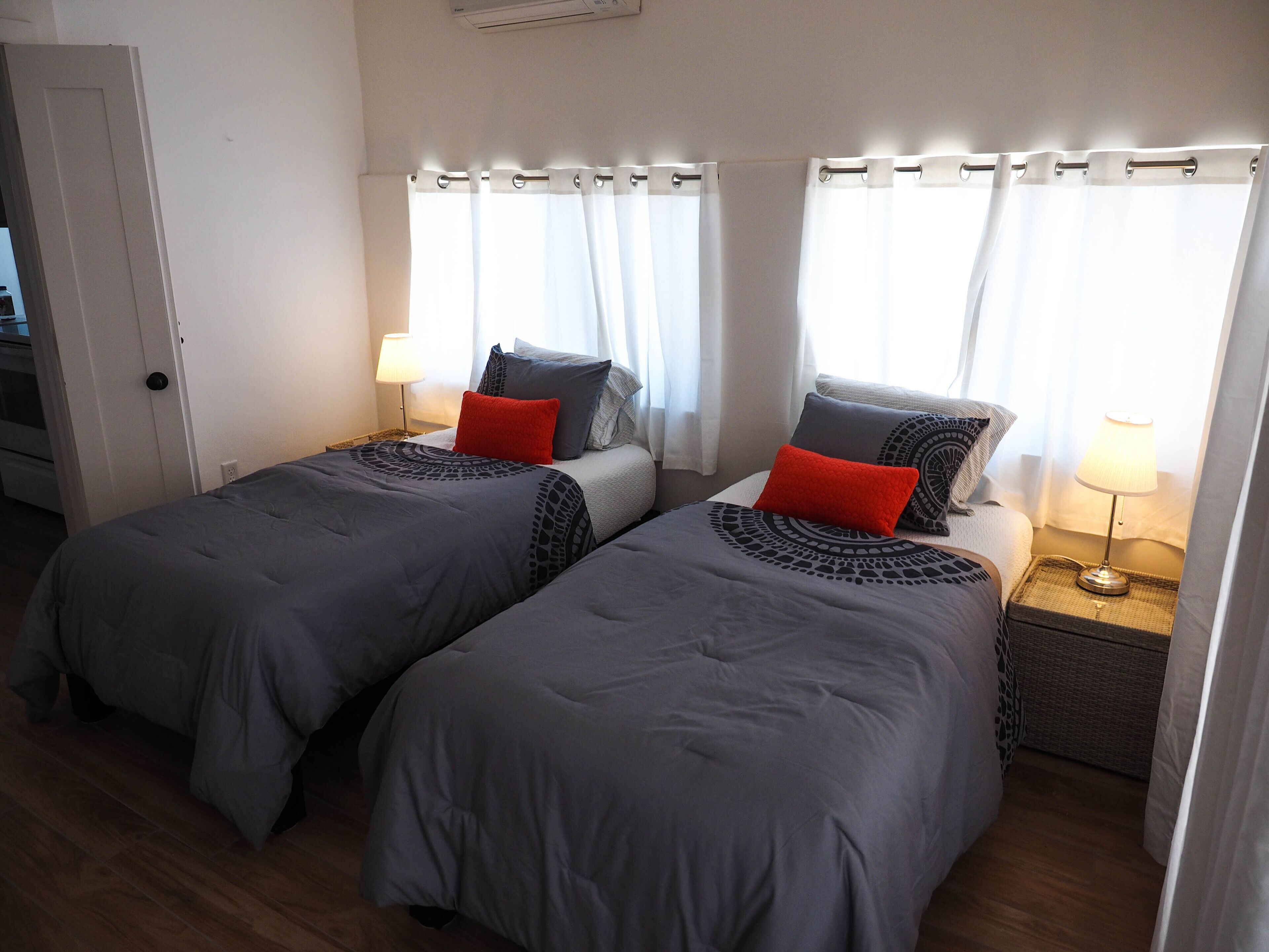 A double room with two beds, each with grey bedding and red pillows, a bedside table with a lamp, and a window with white curtains.