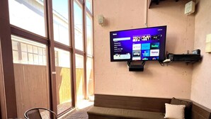 A room with a large flat-screen TV mounted on the wall, a brown sofa, two small stools, wicker chairs, and a window with a view of a building. - Marfa House El Balcon, in the heart of Marfa with direct balcony access (Marfa)