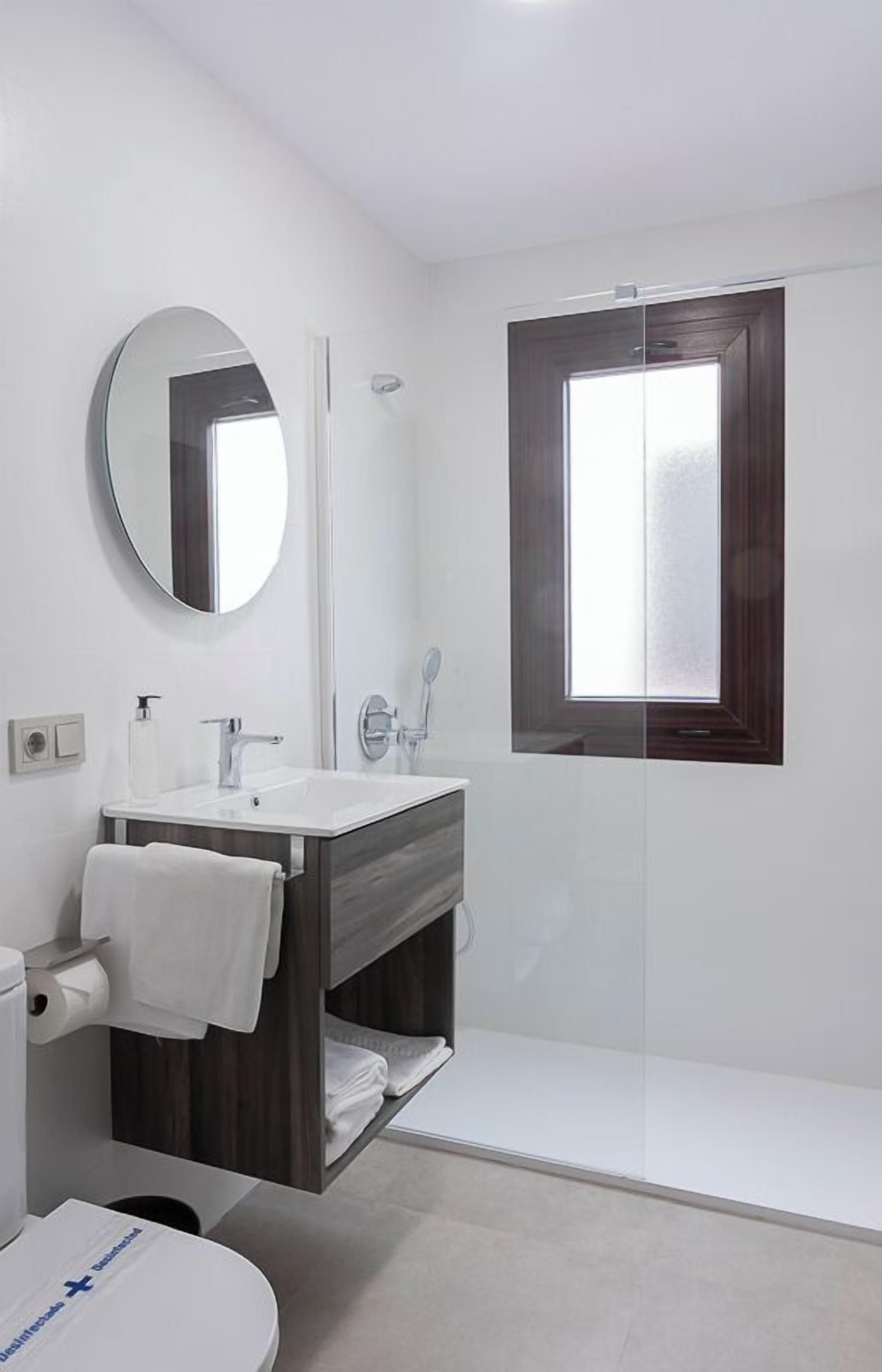 A modern bathroom with a white sink, mirror, and shower. There is a window above the shower and a towel rack with towels.