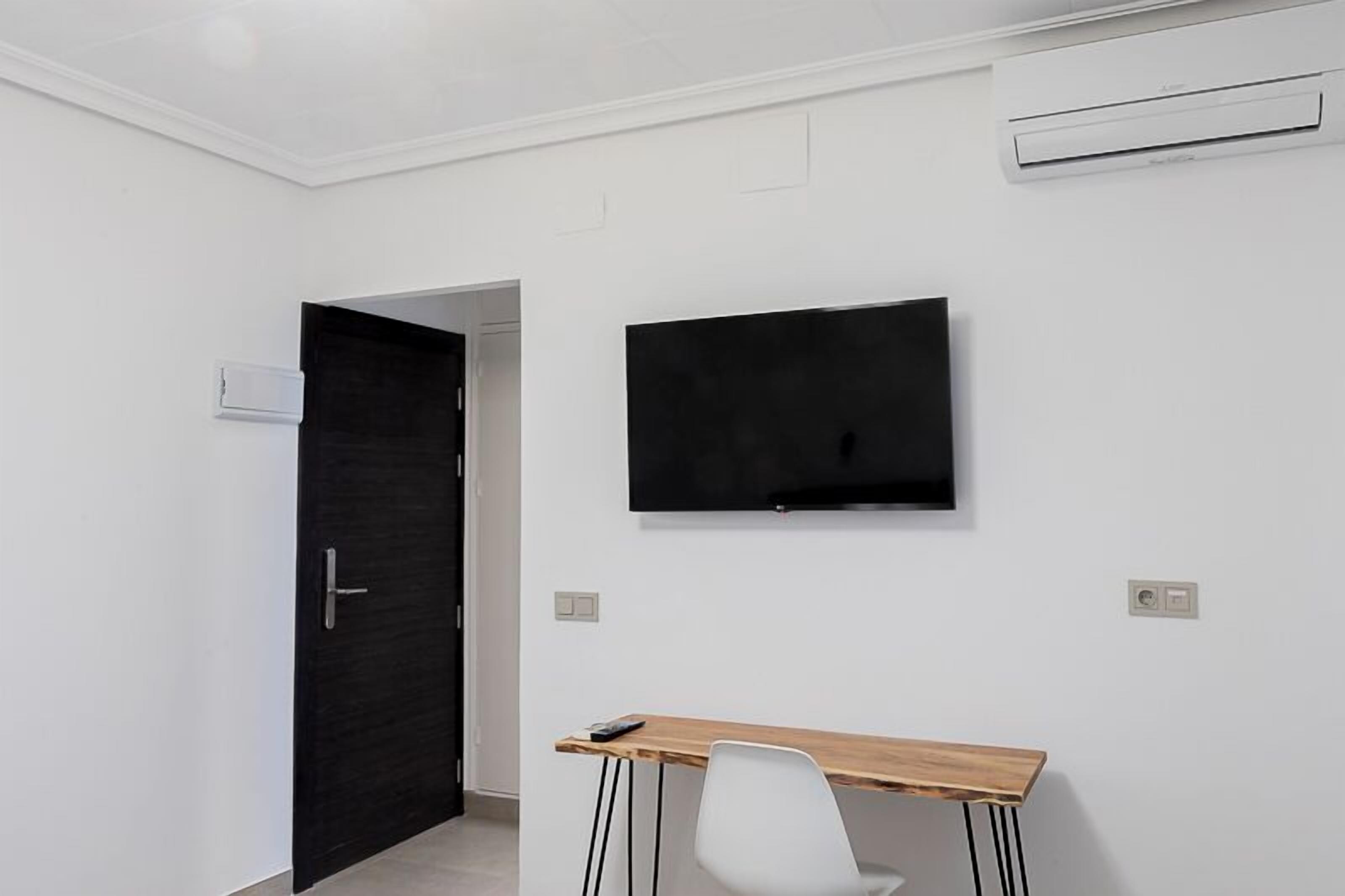 Une chambre moderne dotée d’un téléviseur à écran plat fixé au mur, d’un bureau en bois avec une chaise et d’une porte sombre.