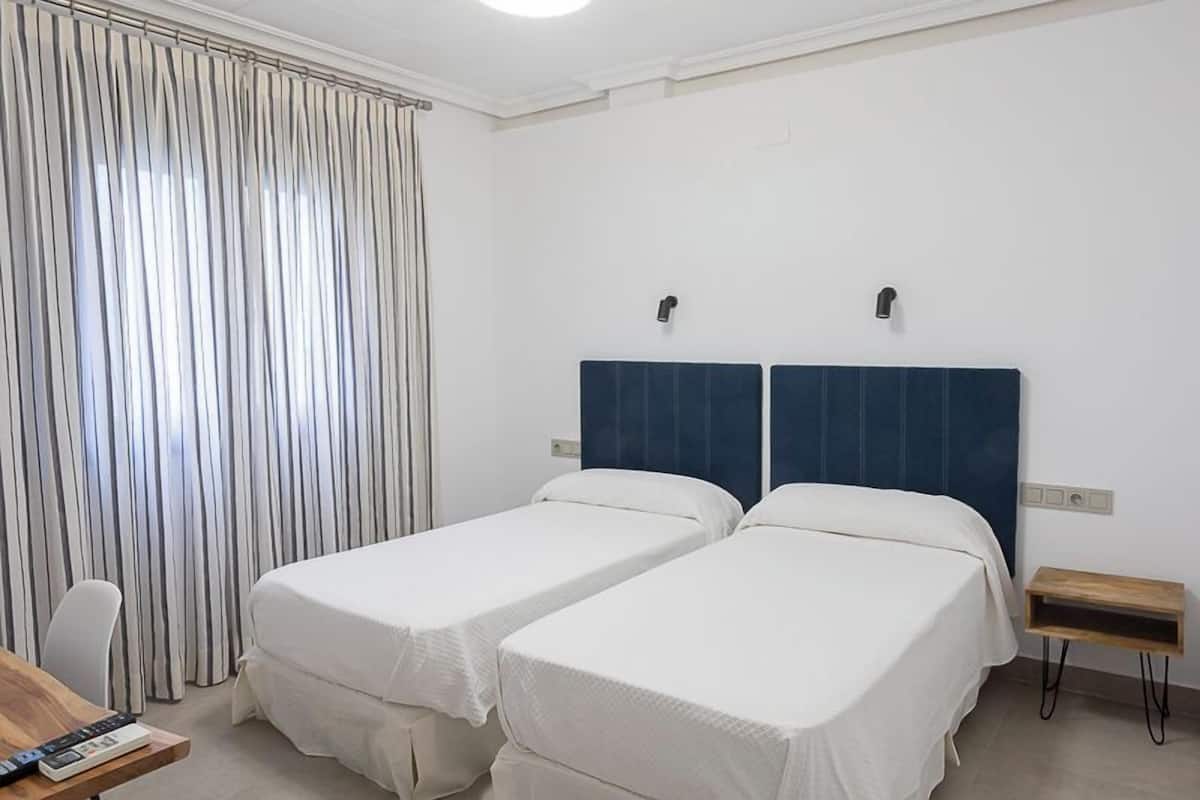 Quarto de hotel com duas camas de solteiro, uma escrivaninha com cadeira e uma janela com cortinas.