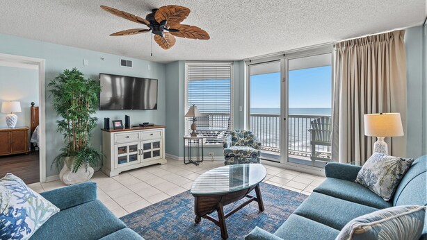 A spacious living room with a large TV, comfortable seating, a ceiling fan, and a view of the ocean through the sliding glass doors.