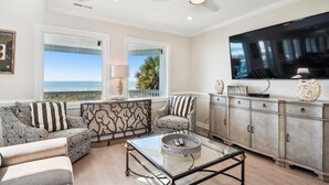 A living room with a large TV, a glass coffee table, and a view of the ocean.