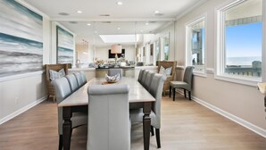 A spacious dining area with a long table and comfortable seating, large windows offering a view of the ocean, and a modern kitchen in the background.