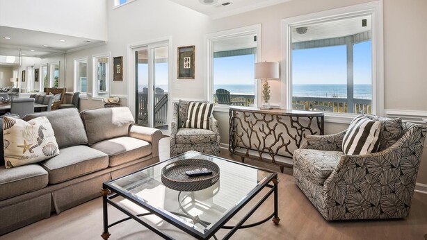 A spacious living room with a large glass coffee table, two patterned armchairs, a sofa, and a dining area with a view of the ocean.
