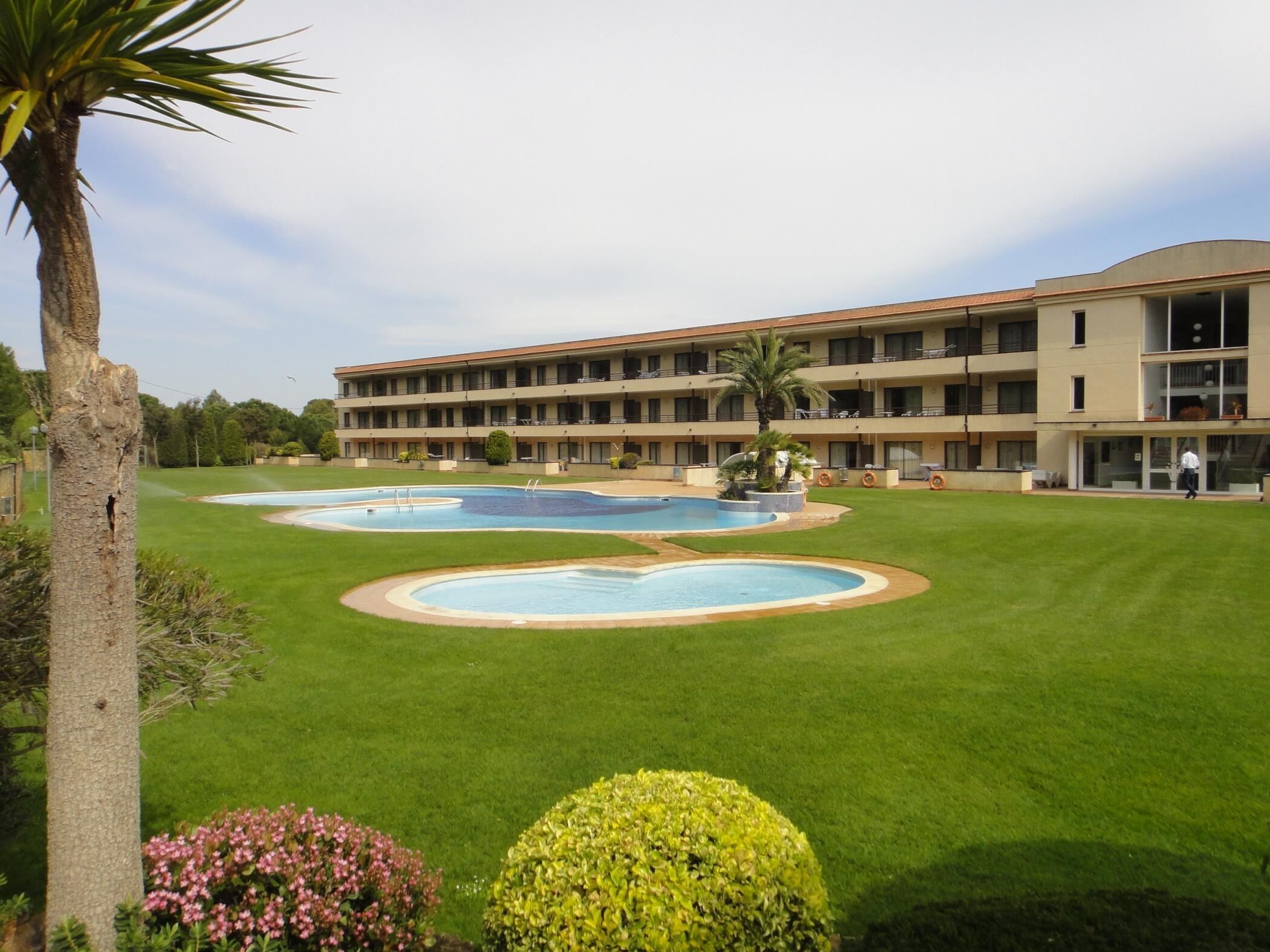 Un hotel con un amplio espacio de piscina, césped bien cuidado y un edificio de varios pisos con balcones.