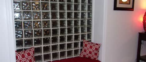Assento junto à janela com almofada vermelha e almofadas estampadas, um quadro na parede e um abajur vermelho sobre uma mesa.