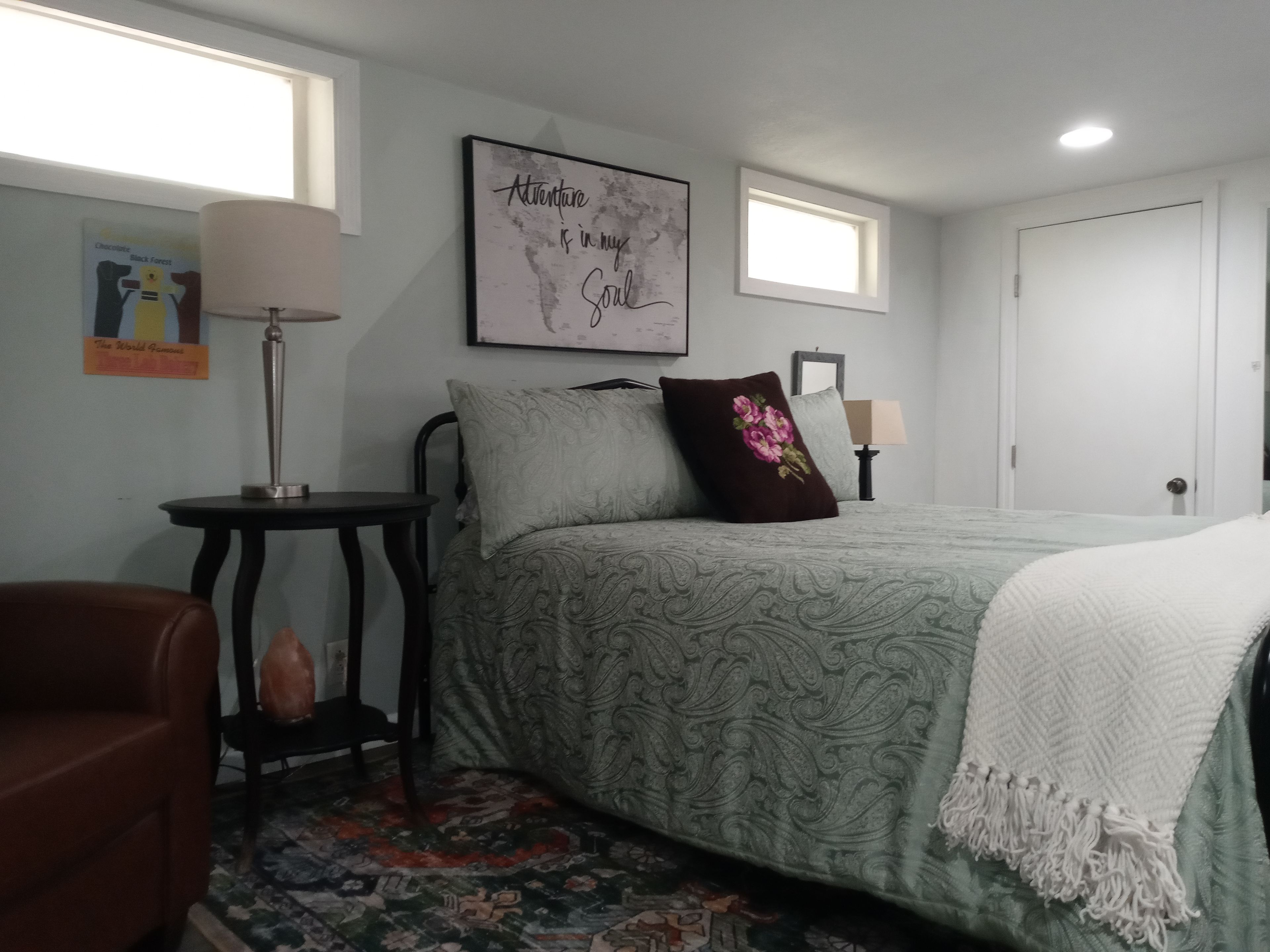A bedroom with a bed, a nightstand, a chair, a window, and a wall with a framed quote.