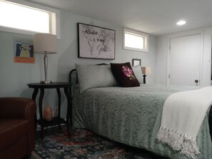 Une chambre à coucher comprenant un lit, une table de chevet, une chaise, une fenêtre et un mur orné d’un cadre avec une citation.