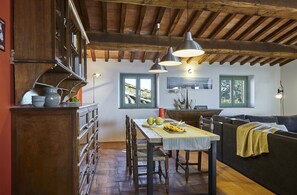 A rustic kitchen with wooden beams, a dining table with chairs, a sofa, and a window with a view of trees. - LA TORRE, AN EAGLE NEST ABOVE THE TUSCANY LANDSCAPE (Castelnuovo Berardenga)