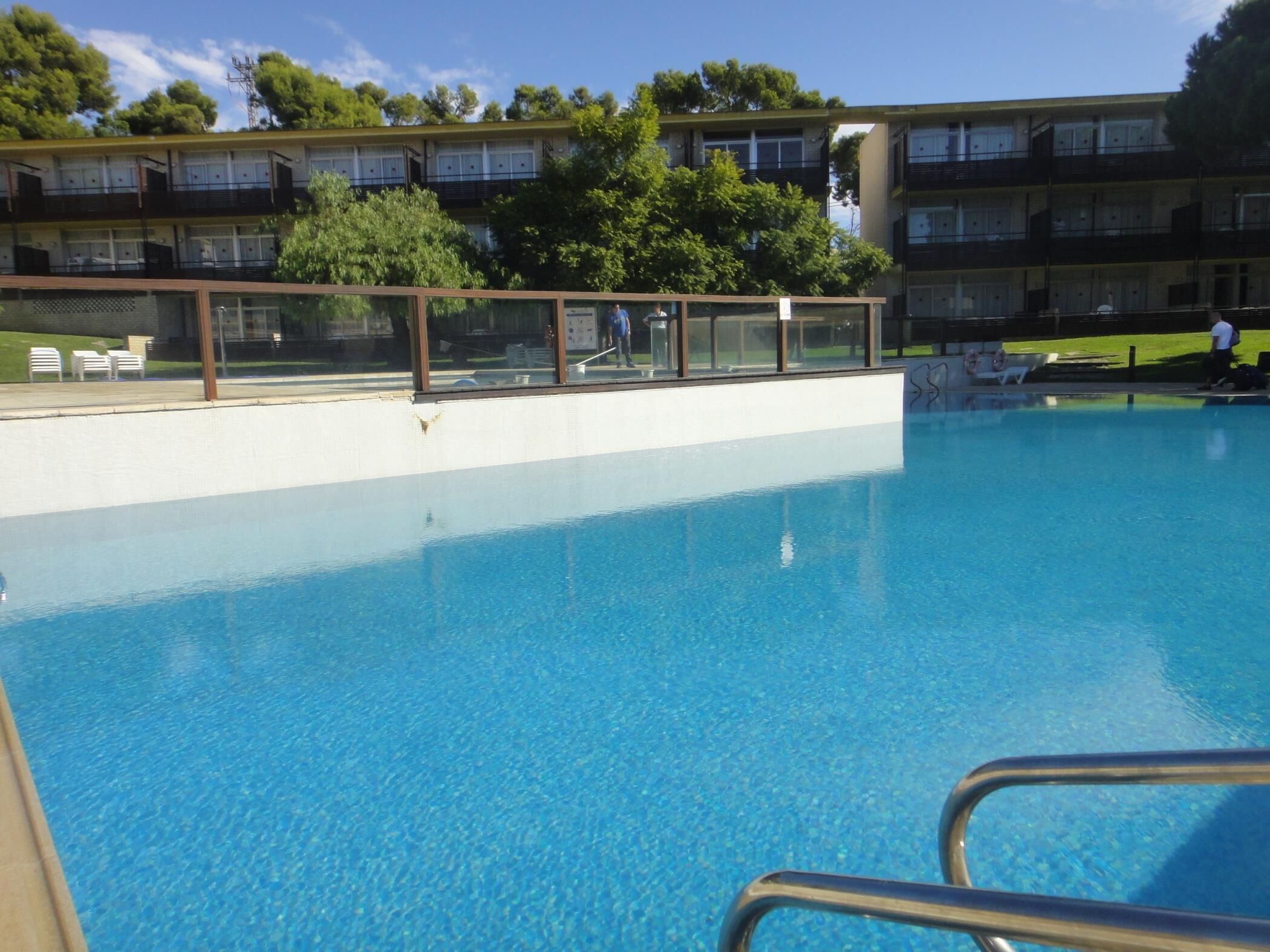 En swimmingpool med kristallklart blått vatten, omgiven av ett glasräcke. I bakgrunden syns höga byggnader med balkonger och en gräsyta.