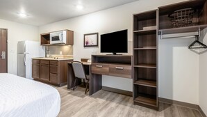 Una habitación de hotel funcional con kitchenette, una cama, un escritorio con silla, televisor y armario con estantes.