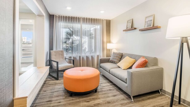 Una sala de estar moderna con un sofá gris, un reposapiés naranja, una silla y un ventanal con cortinas ligeras.