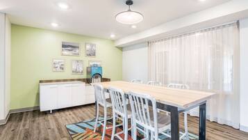 A dining area with a wooden table and white chairs, a kitchen with white cabinets, and a window with sheer curtains.