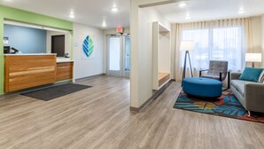 Un vestíbulo de hotel con mostrador de recepción, un área de espera con sofá y reposapiés, y un ventanal con cortinas.