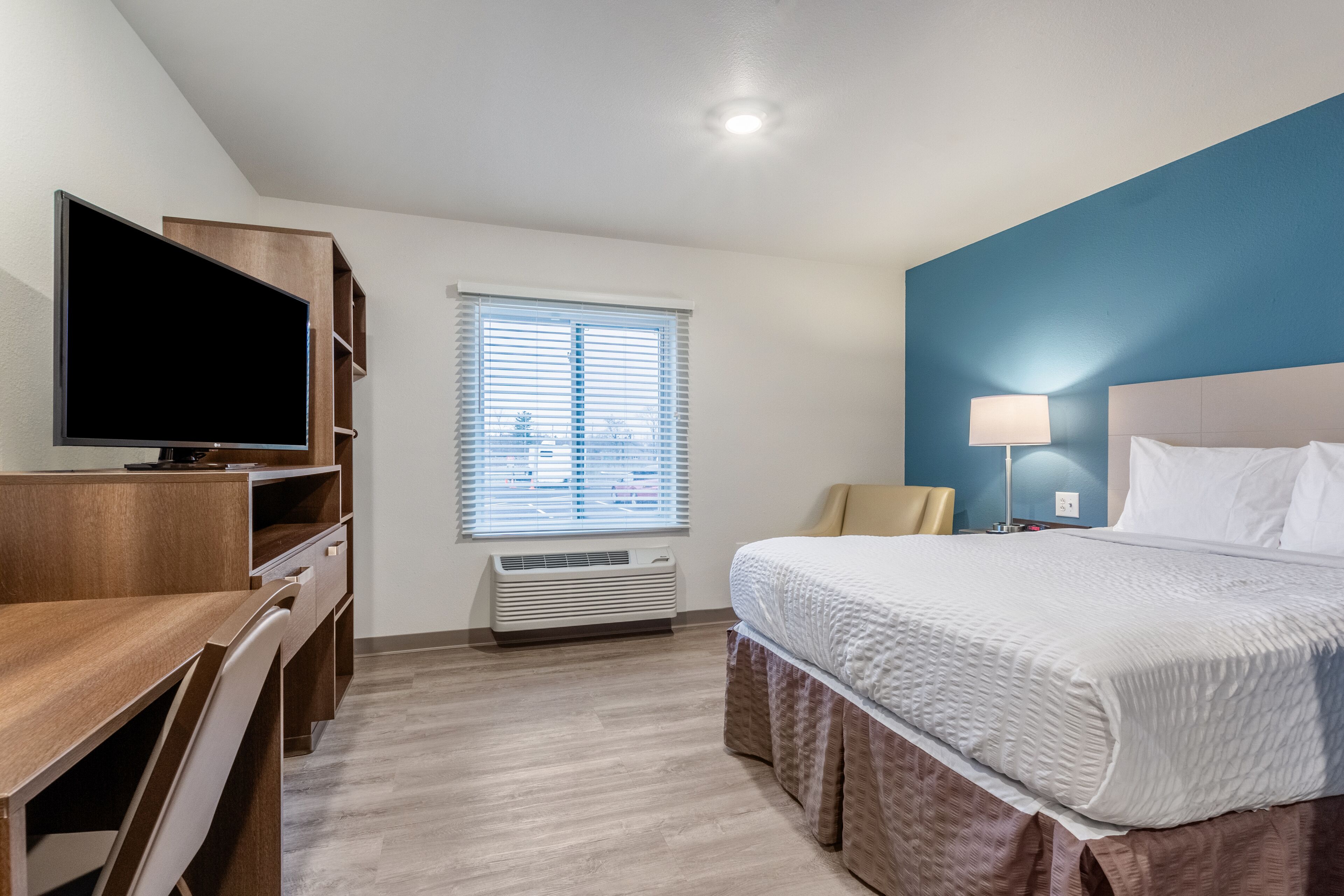 A hotel room with a bed, a desk, a television, a window with blinds, and a lamp.
