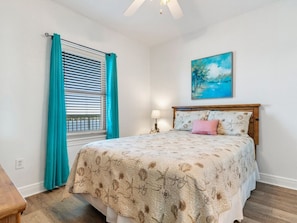 A bedroom with a bed, a window with curtains, a painting on the wall, and a nightstand with a lamp.