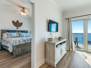 A bedroom with a bed, a ceiling fan, a TV, a small table with a basket, and a view of the ocean through a sliding glass door.