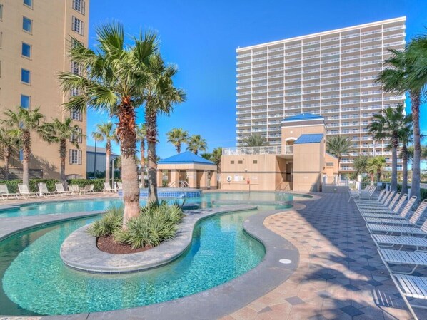 A hotel with a swimming pool, palm trees, and a large building in the background.