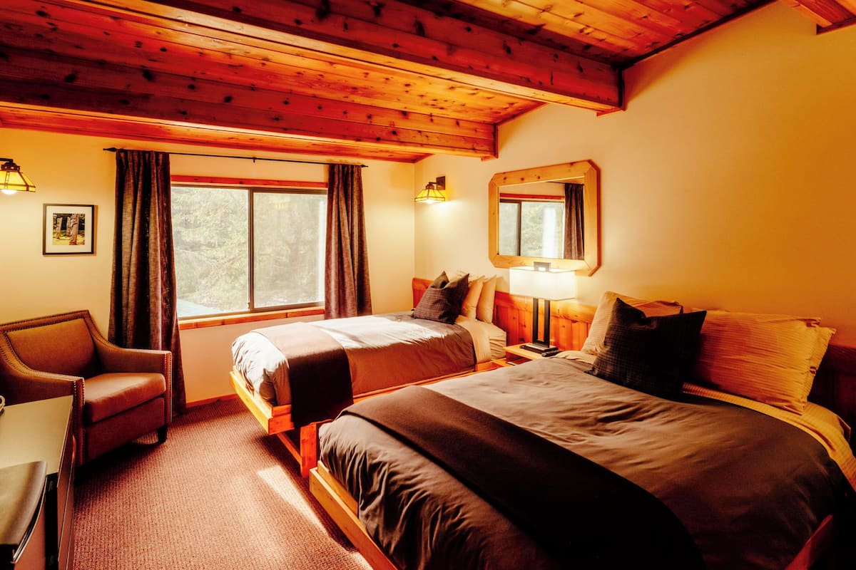 A room with a wooden ceiling, a bed with a headboard, a desk, a chair, a window with curtains, and a wall-mounted mirror.