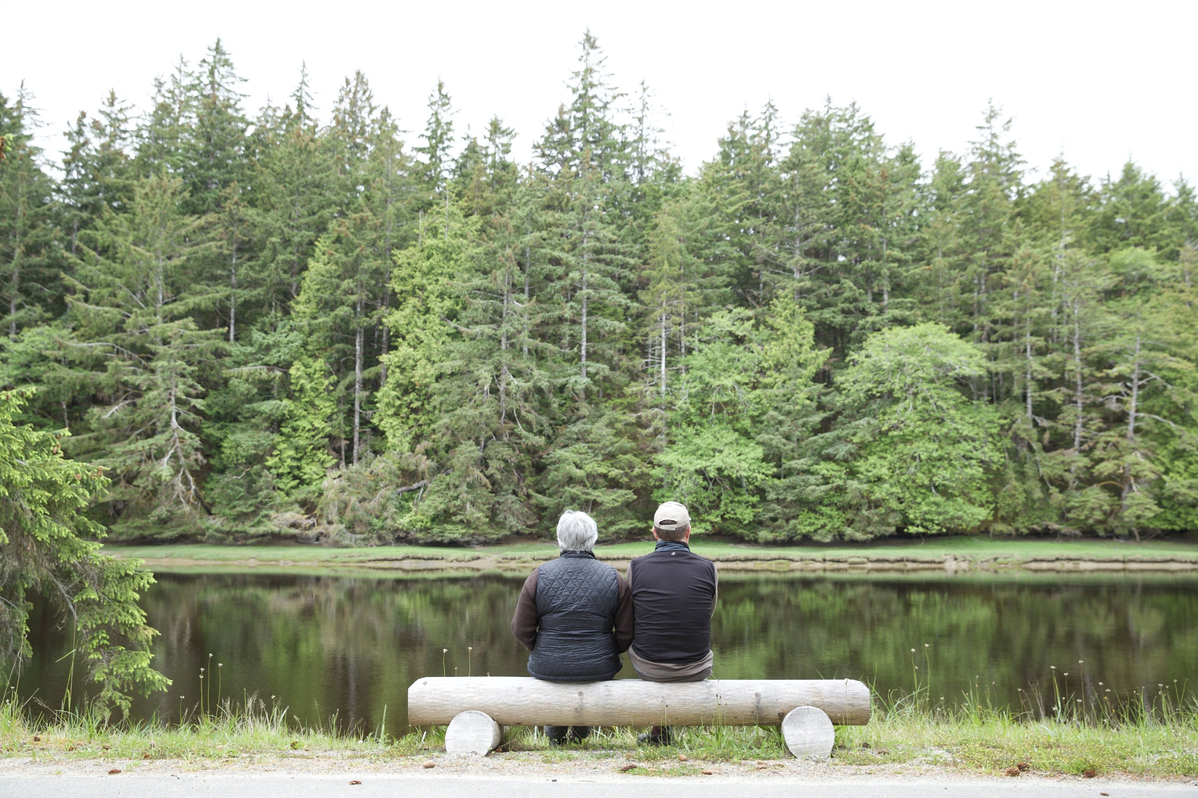 Two people sitting on a bench by a lake with a forest in the background.