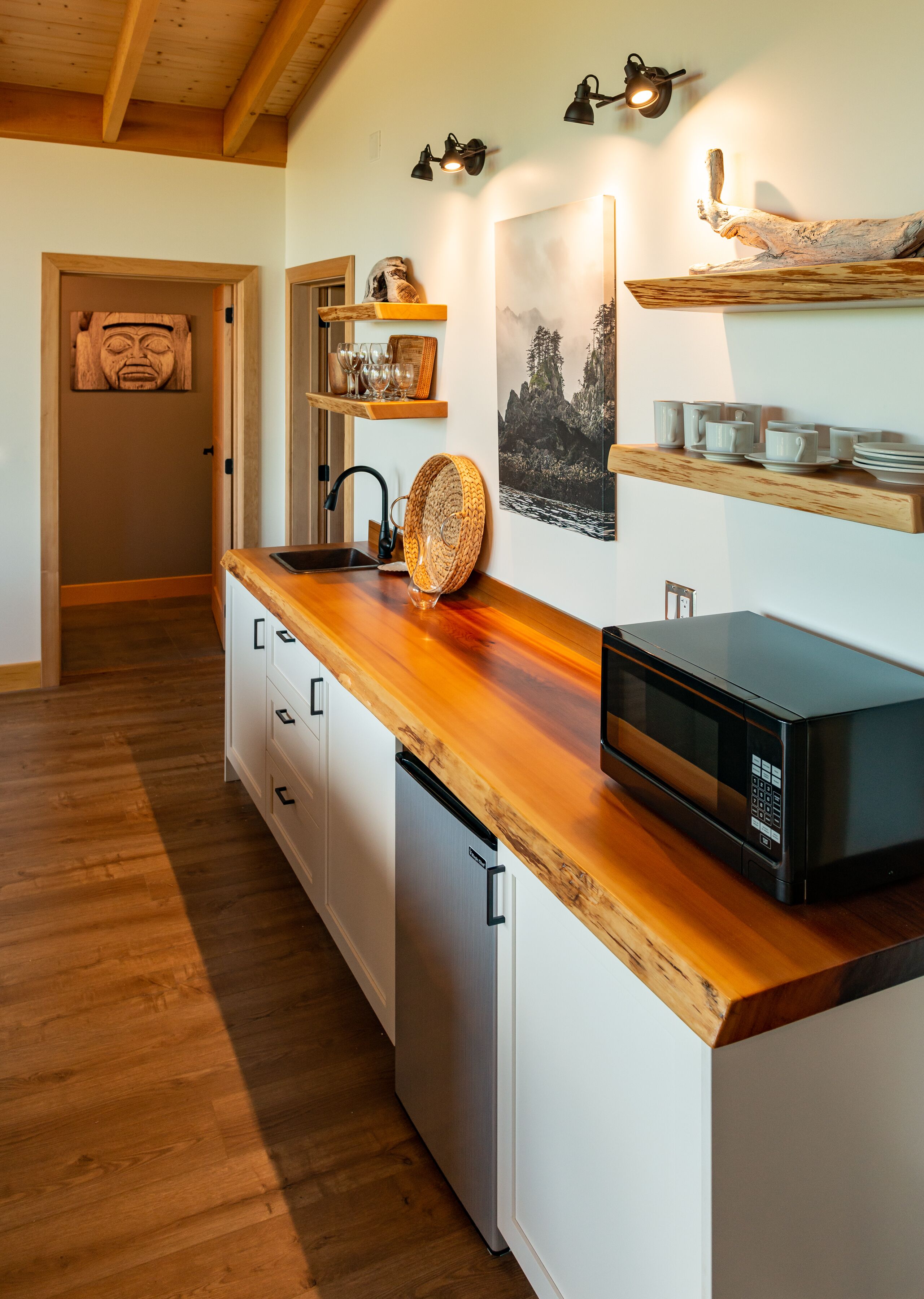 Een moderne keuken met houten aanrecht, een ingebouwde vaatwasser en een magnetron. Er zijn planken met kopjes en een schilderij van een berglandschap.