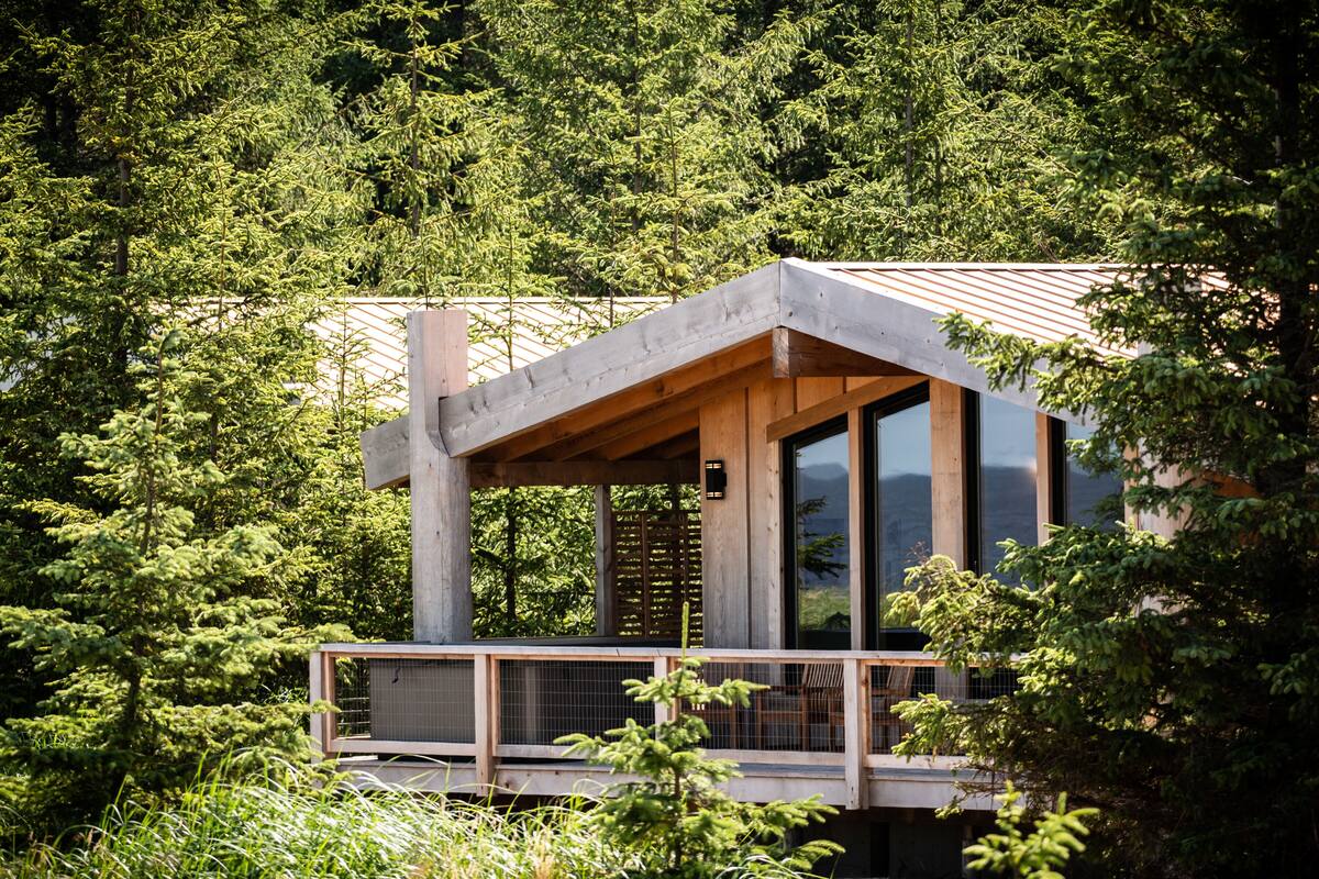 A modern cabin with a wooden deck and large windows, surrounded by dense forest.