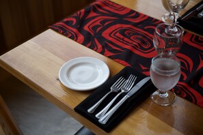A wooden table with a black and red patterned tablecloth, a white plate, a black napkin with silverware, and a glass of a light-colored beverage.